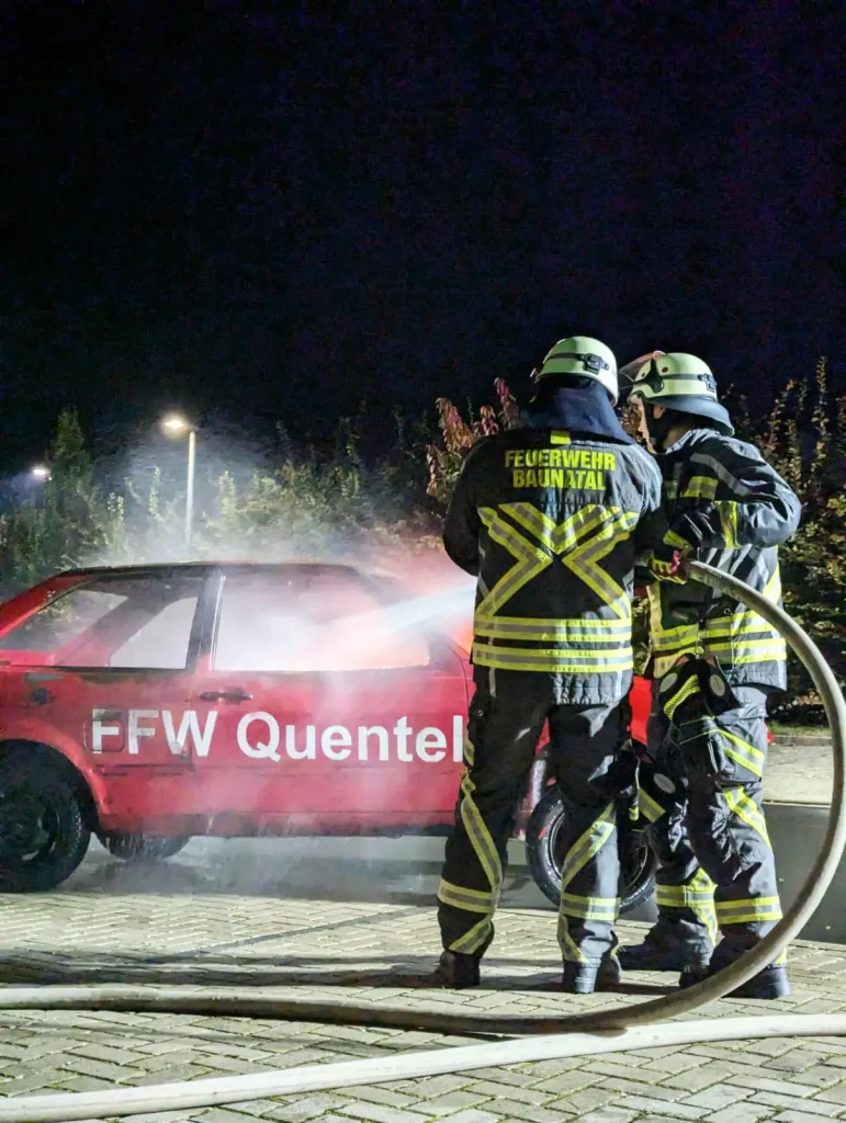 Zwei Feuerwehrleute löschen ein brennendes Übungsauto.